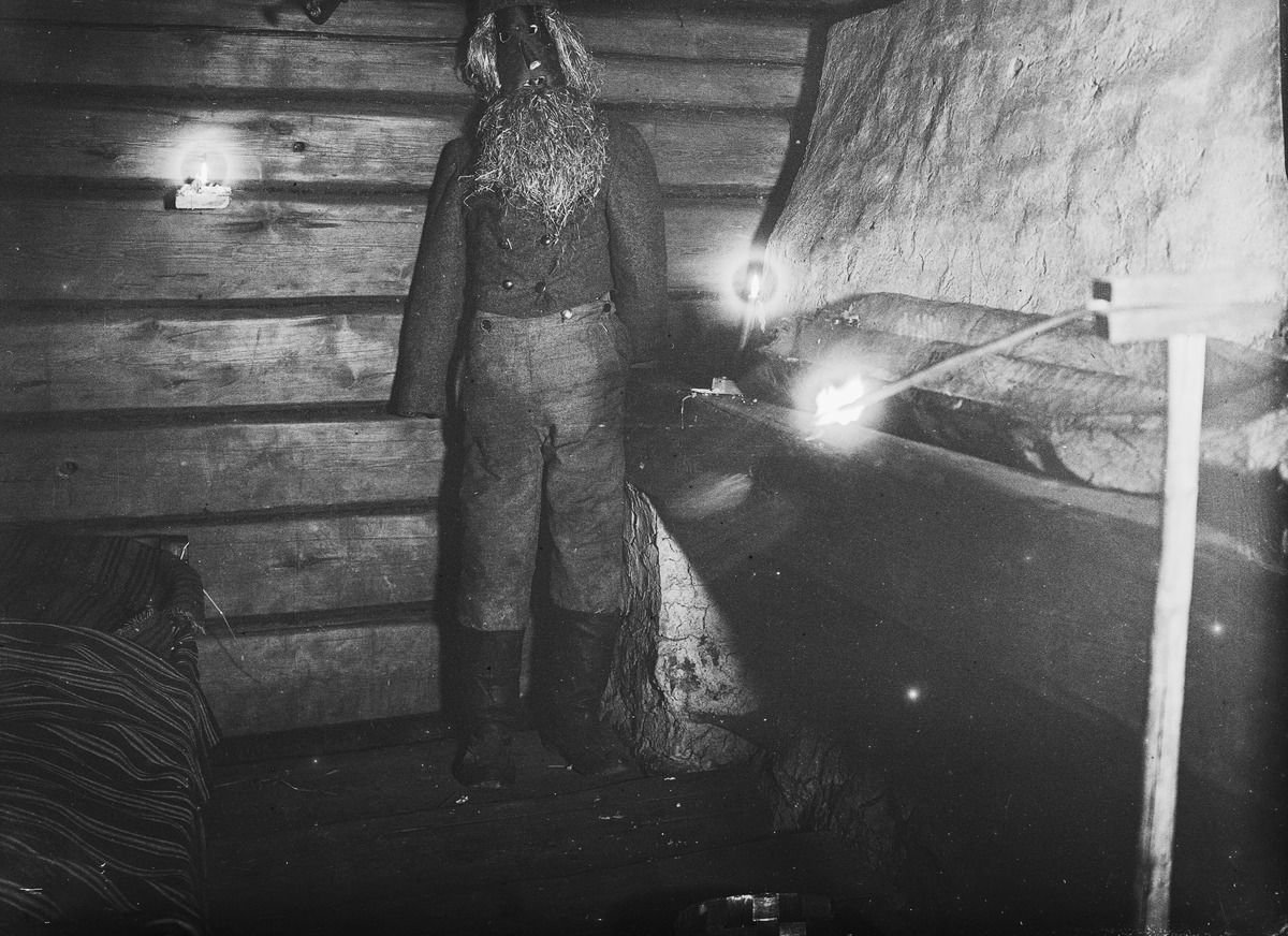 A life-size dressed figure standing beside the stove in a dim log cabin.