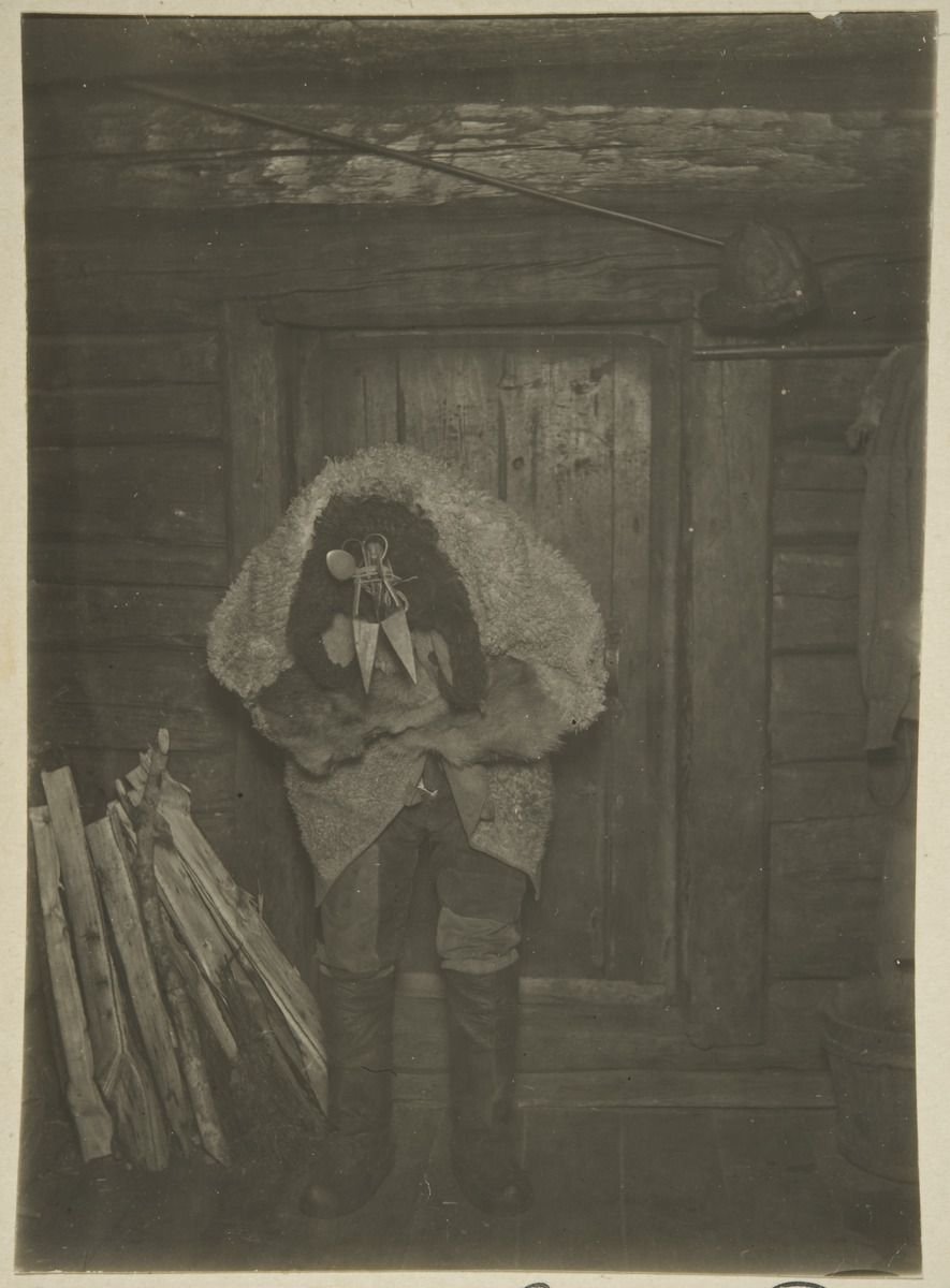 A figure in a pale hooded costume with a mask, standing by the door of a log building.