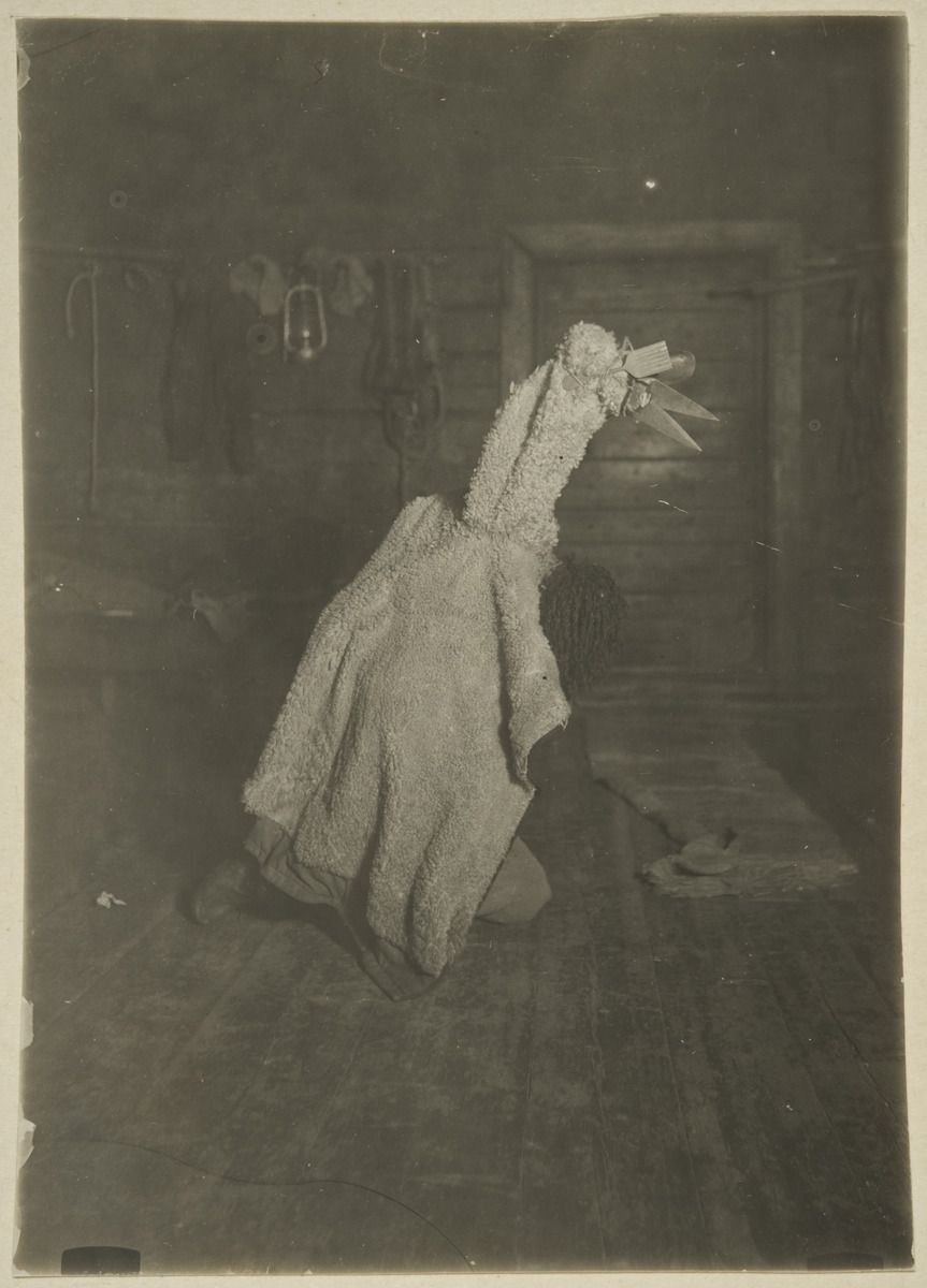 A figure covered in a long pale cloth costume, bent forward inside a log room.