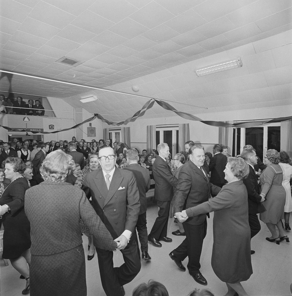 Couples dancing in a crowded hall decorated with paper garlands.