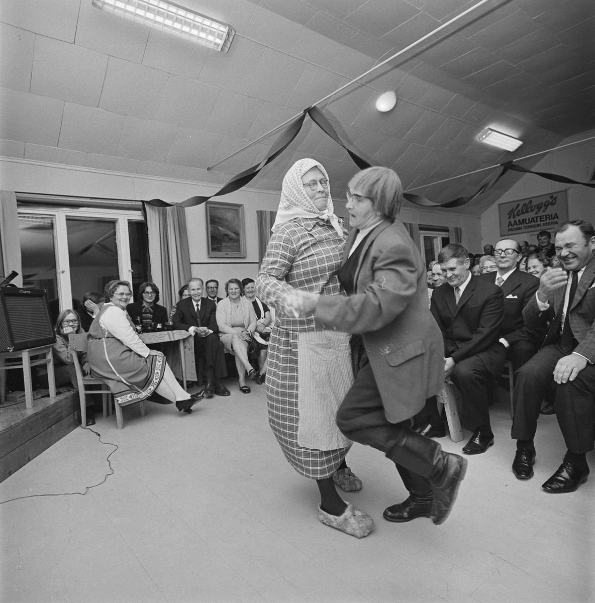 Two performers in folk costume dancing in front of a seated audience.