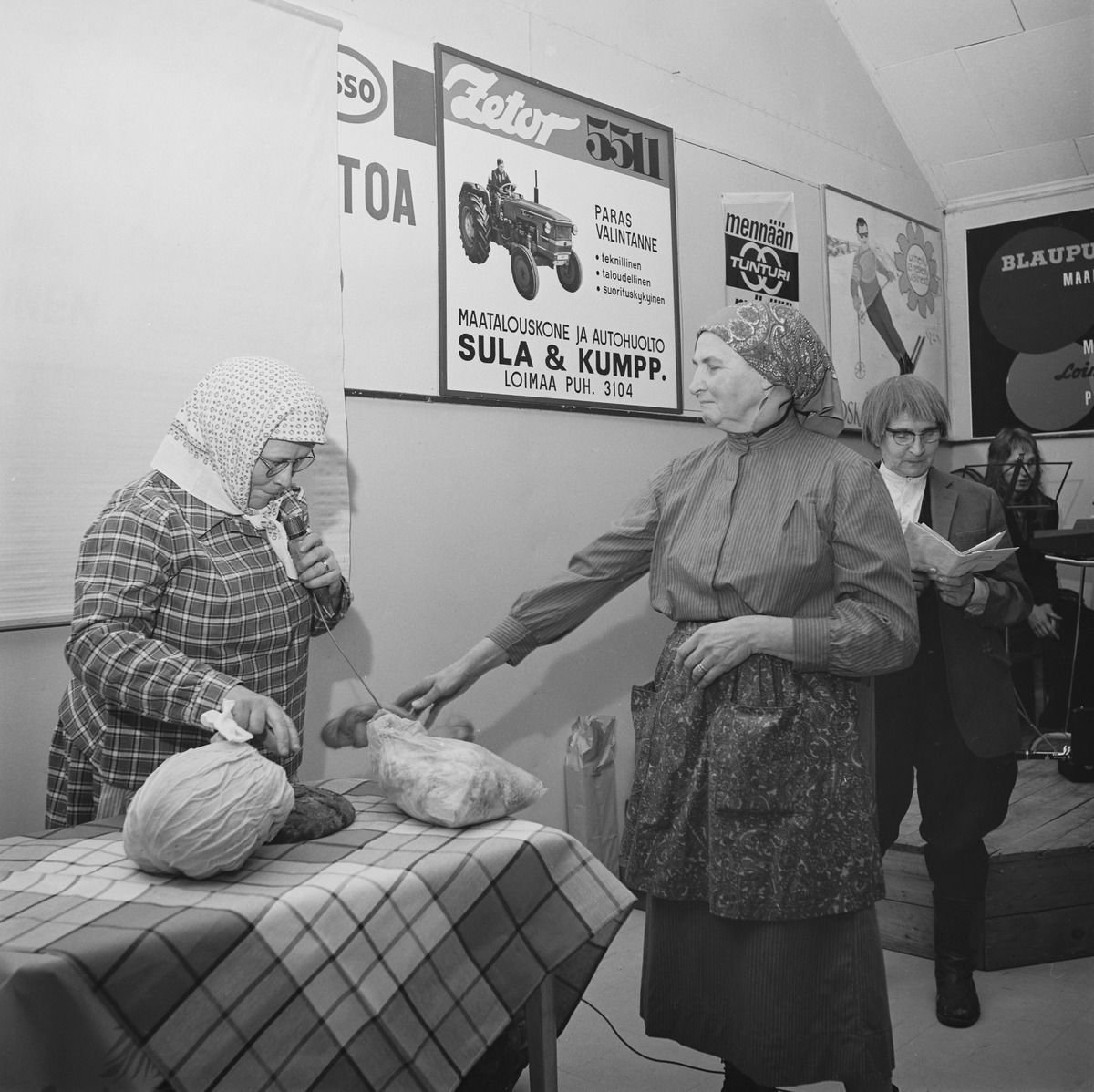 Women in headscarves and aprons acting a scene beside a table with a large round vegetable and loaves, in a decorated hall.