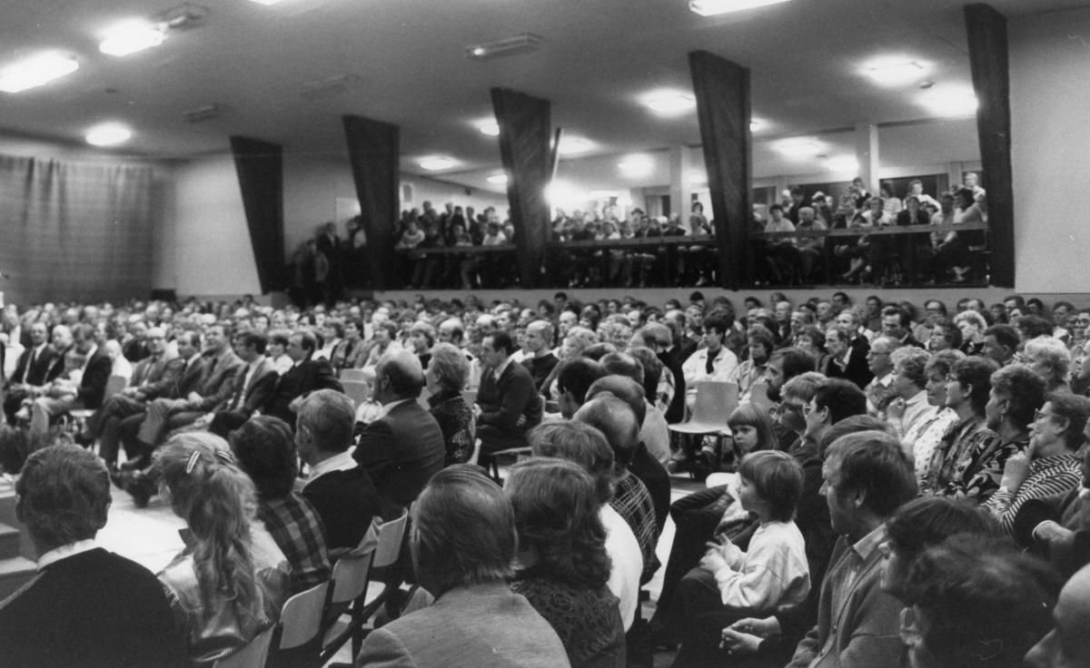 A large audience seated in a long hall, seen from the back towards the stage.