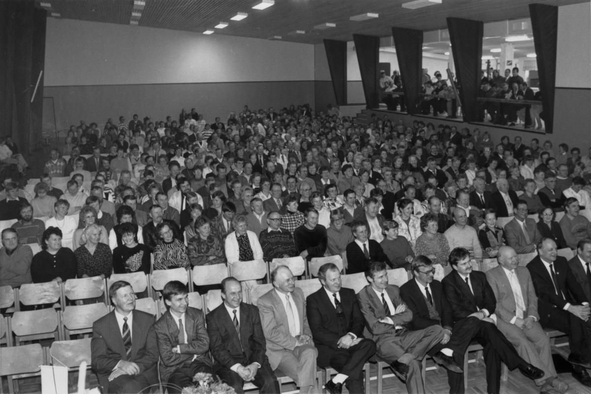 Rows of people seated in a hall, men in suits in the front row.